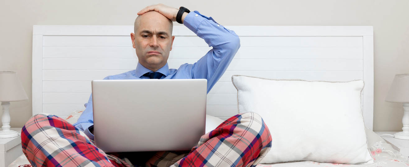 Man sitting up in bed, using a laptop and wearing a shirt and tie with pyjama trousers, looking unhappy.